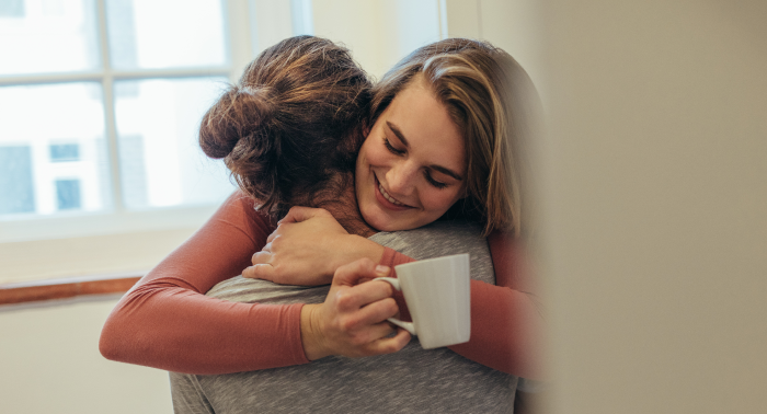 Two women hugging whilst one it holding a white mug