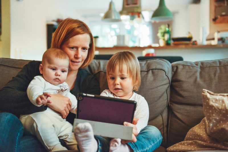 A mother is sitting on a couch with her two young children, engaging with a tablet. The toddler on her left is holding the tablet, focused on the screen, while the baby in her lap gazes at it curiously. The family appears to be in a cozy living room with warm lighting and a blurred background, suggesting a comfortable and relaxed atmosphere at home.