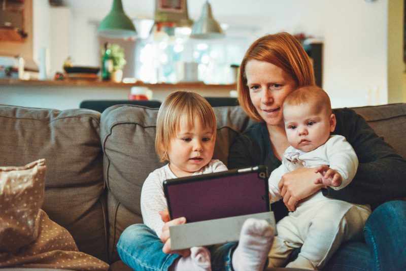 A mother is sitting on a couch with her two young children, engaging with a tablet. The toddler on her left is holding the tablet, focused on the screen, while the baby in her lap gazes at it curiously. The family appears to be in a cozy living room with warm lighting and a blurred background, suggesting a comfortable and relaxed atmosphere at home.