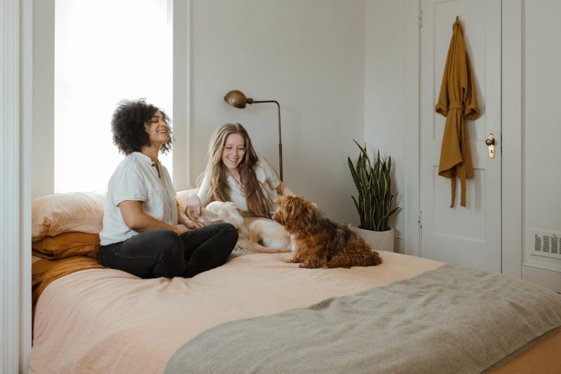 a woman with early curly hair and a woman with long straight hair and glasses sat on bed smiling at a dog