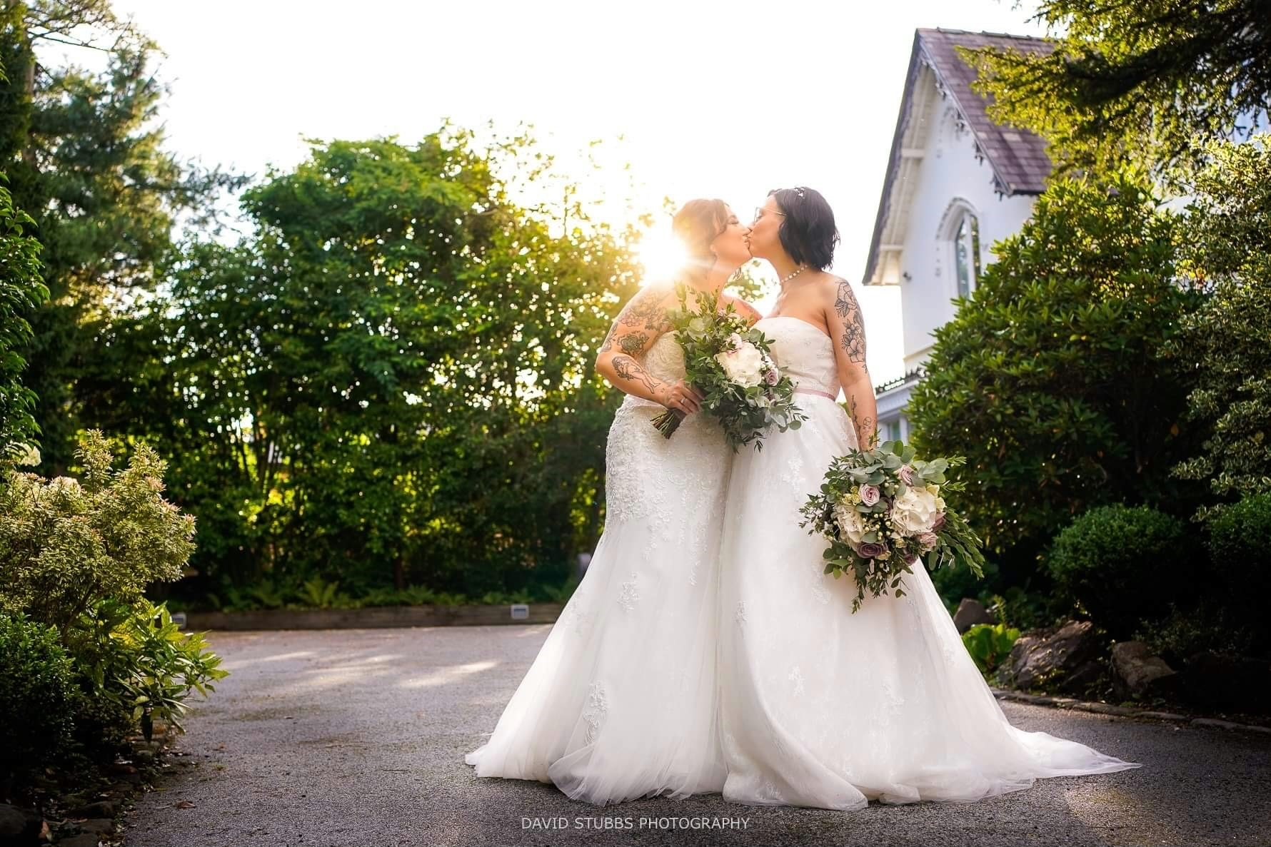 Two women with tattoos in white wedding dresses kissing holding flowers