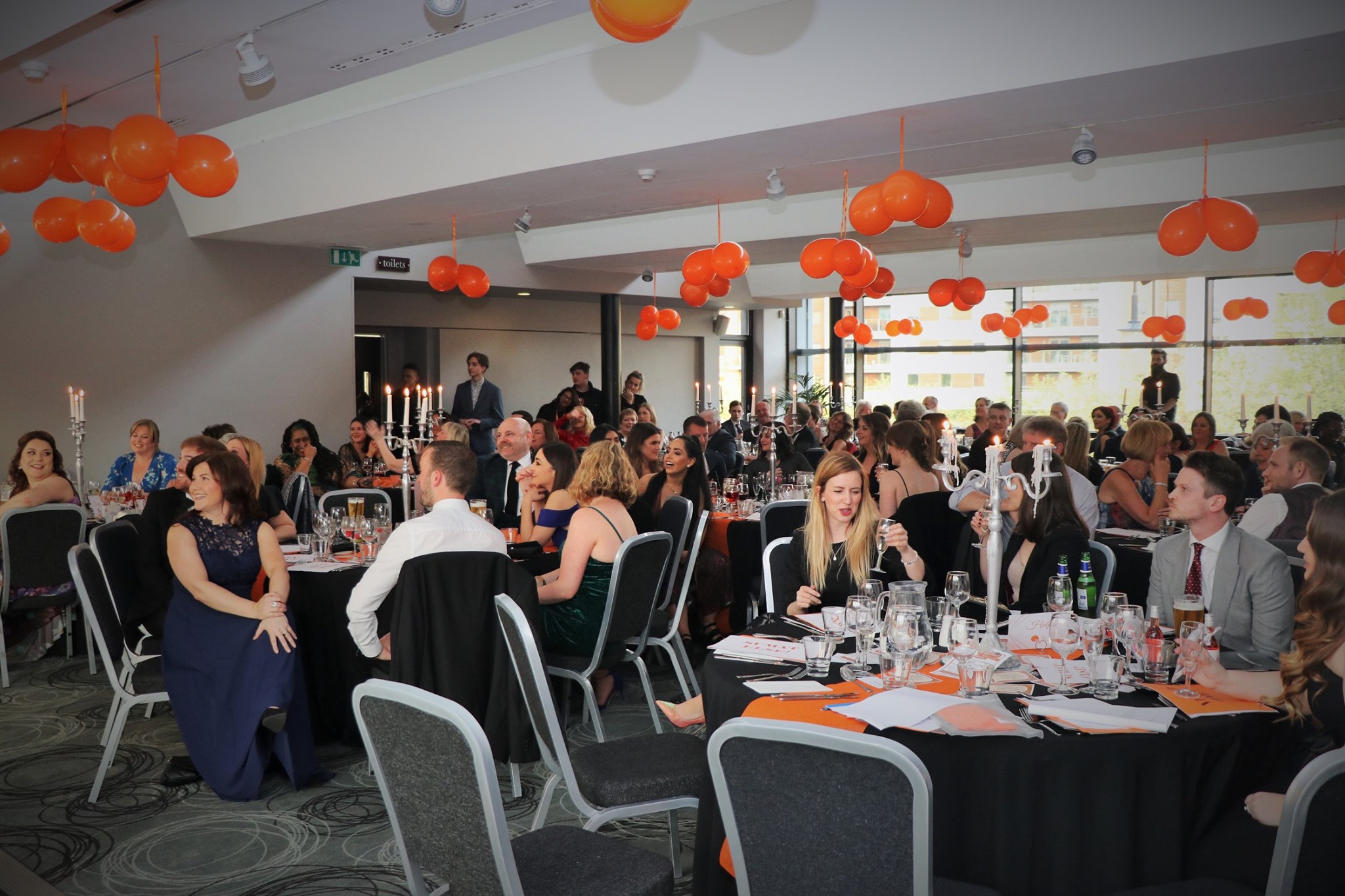 a form party with orange balloons hanging from the ceiling and the room full where people are sat down