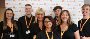 Team of people stand in front of branded event signage. They wear back with highly visible orange TLC lanyards.