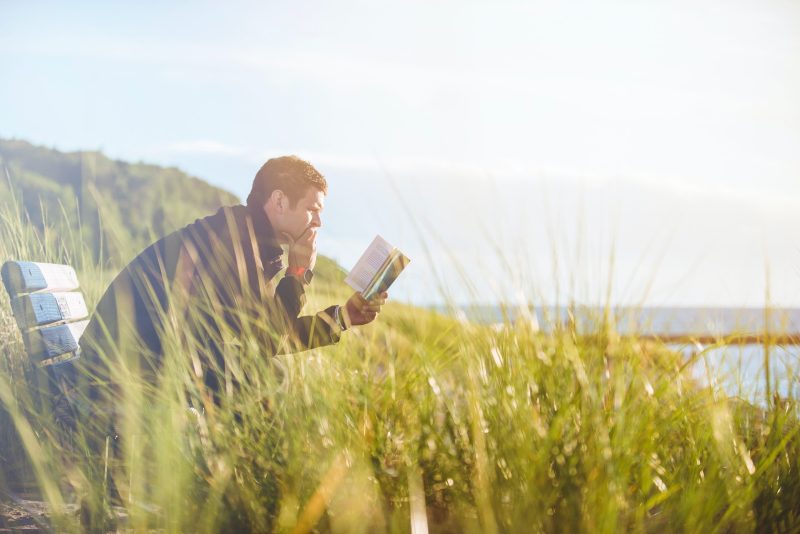 a man sat on a bench surrounded by tall grass reading a book