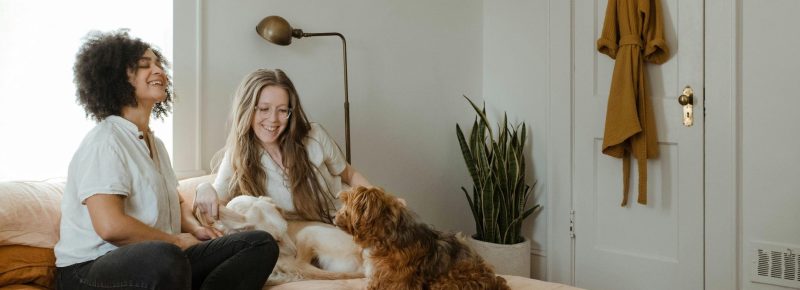a woman with curly hair and a woman with long straight hair and glasses sat on bed smiling at a dog