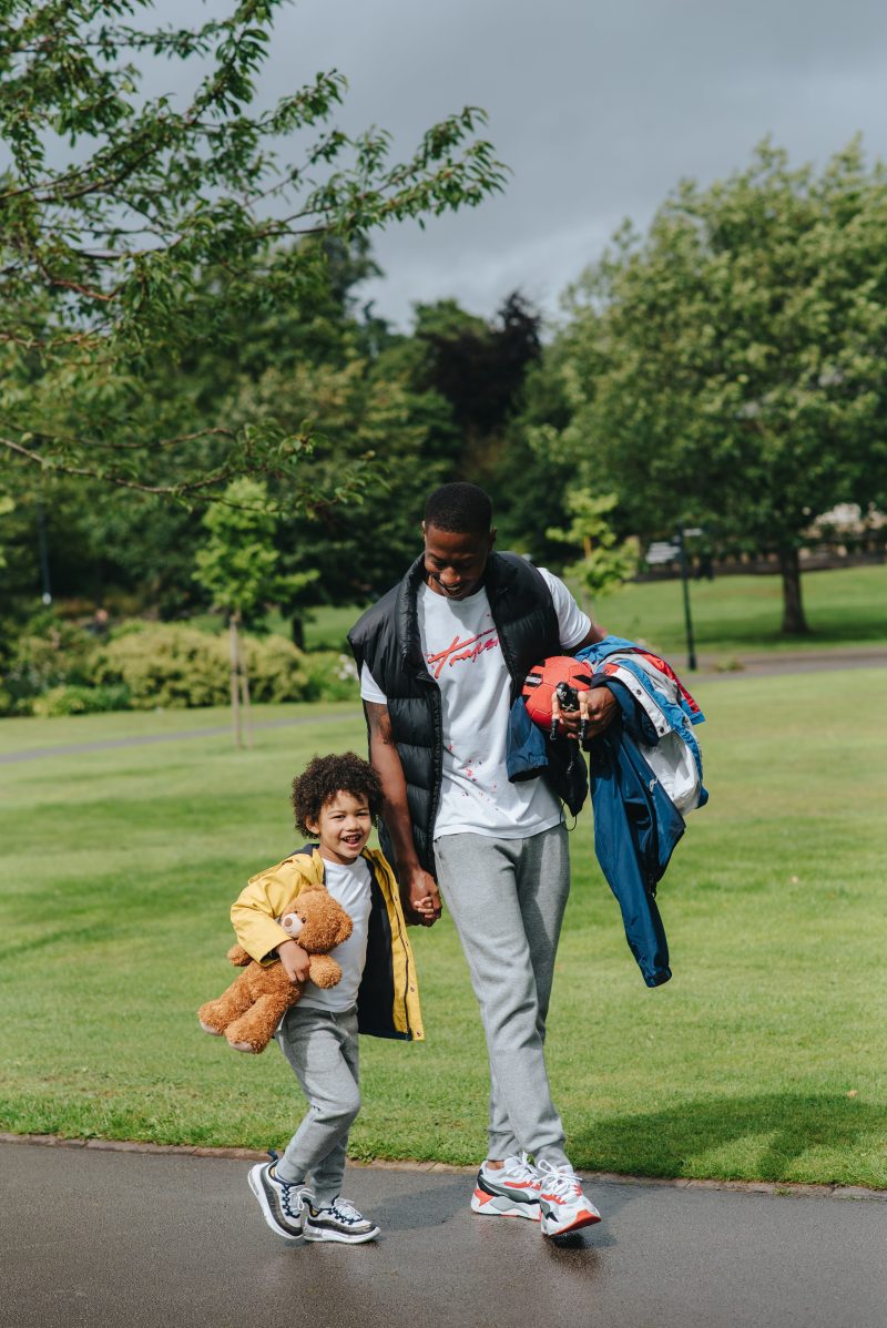 man walking in the park with a football and coat in his arm with his son who's carrying a teddy bear