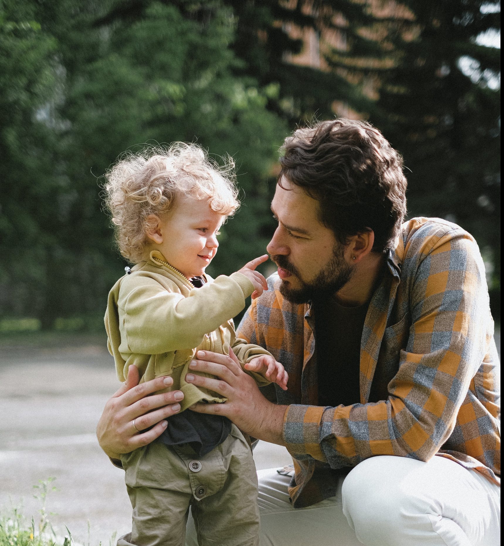 young dad crouching to his son with blonde curly hair touching his nose in the outdoor