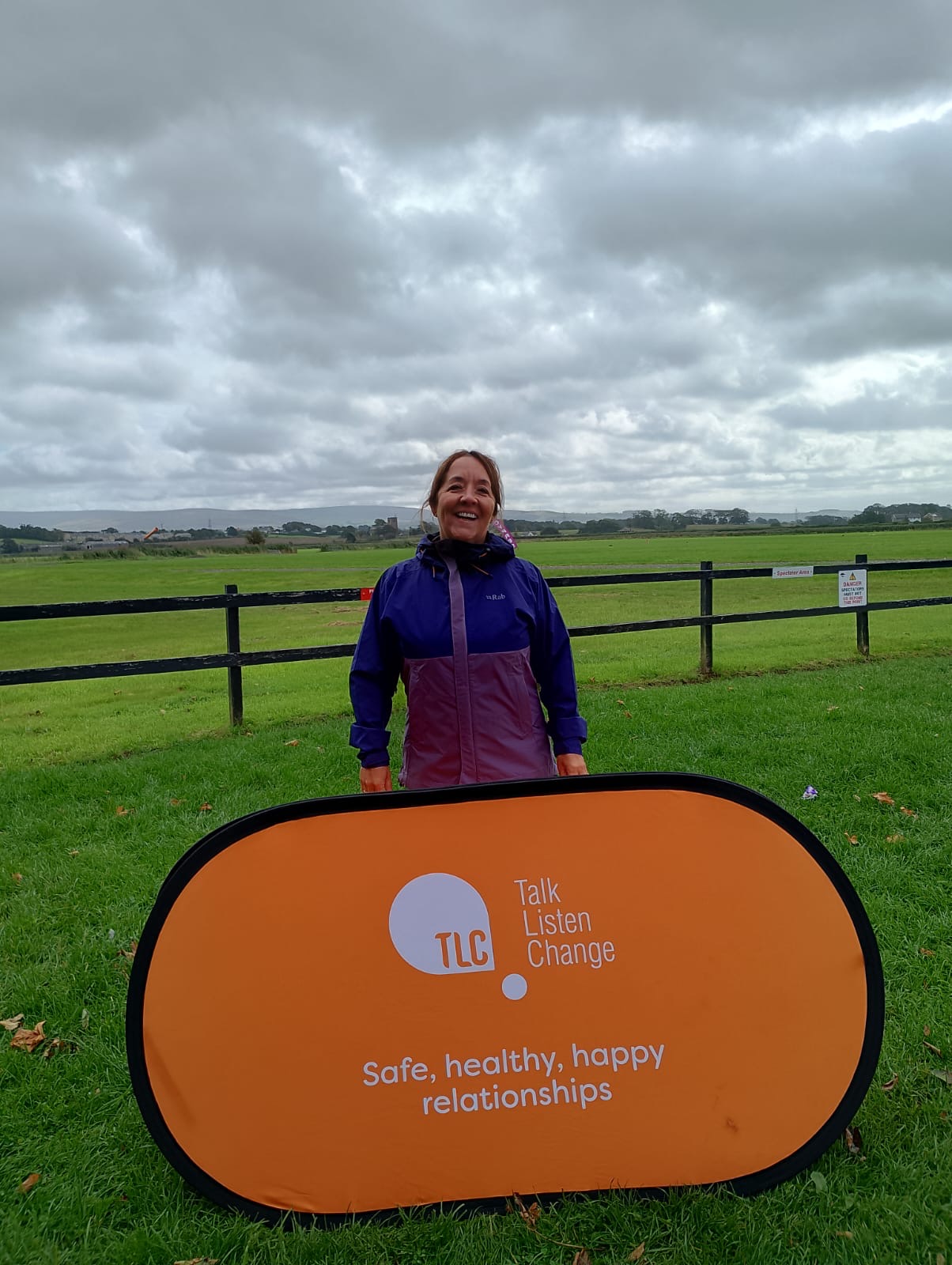 a woman smiling in a purple and blue coat posing in a field with a TLC: Talk, Listen, Change sign in a field