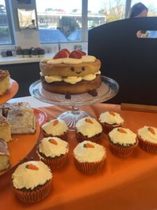 Victoria sponge cake with a smiley face with carrot cake cupcakes in front on an orange table cloth