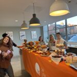 Group of women posing around cake sale display with orange table cloth under the cakes