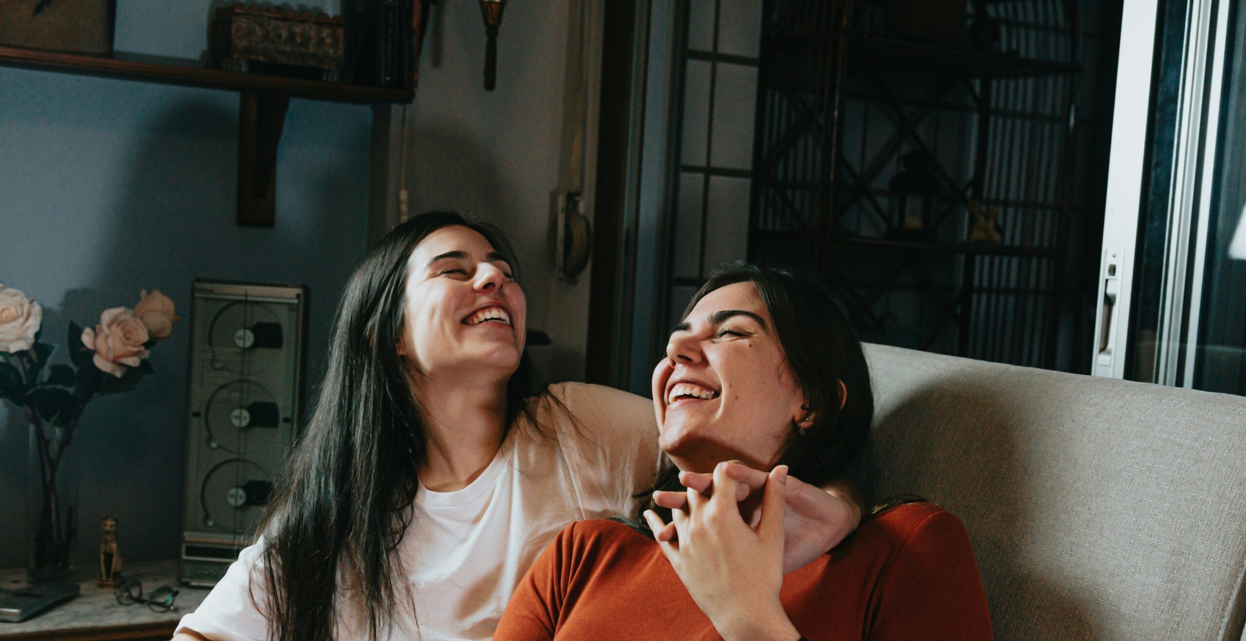 Two woman with dark hair sat on a couch smiling with their hand intertwined