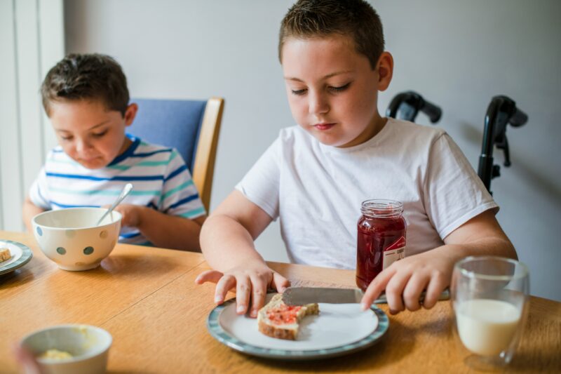 two primary school aged boys sit at the dinner table, both spreading jam onto toast. They are in a home setting as though they are brothers. One of the children is in a wheelchair