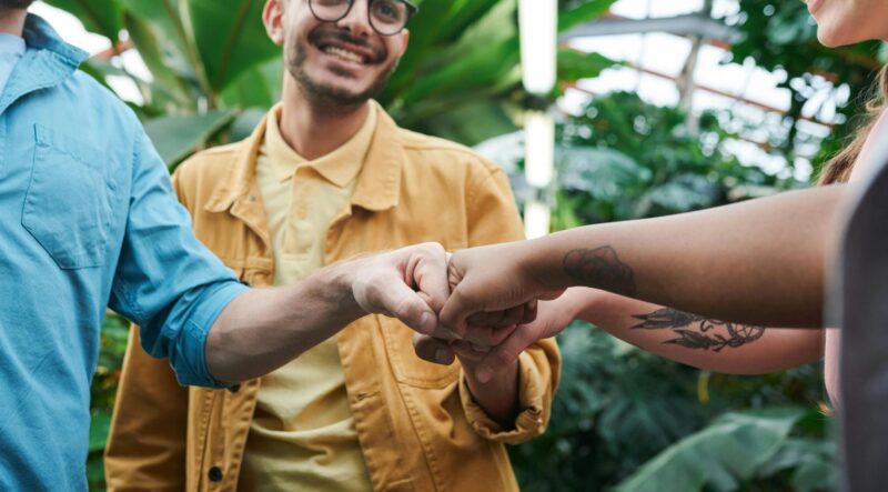 a group of people fist bumping together. Smiling with a backdrop of greenery