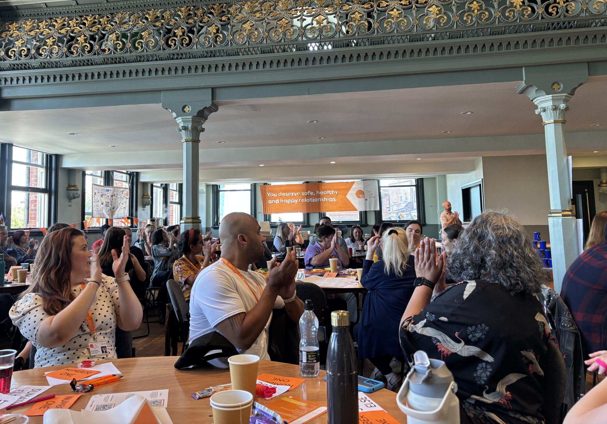 landscape shot of an informal staff conference. People are clapping and in the background is a branded banner reading safe, healthy and happy relationships
