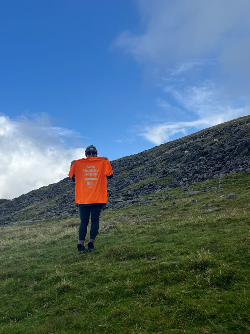 A woman stood on the side of snowdon holding an orange TLC t-shirt saying safe, healthy, happy and a TLC logo.