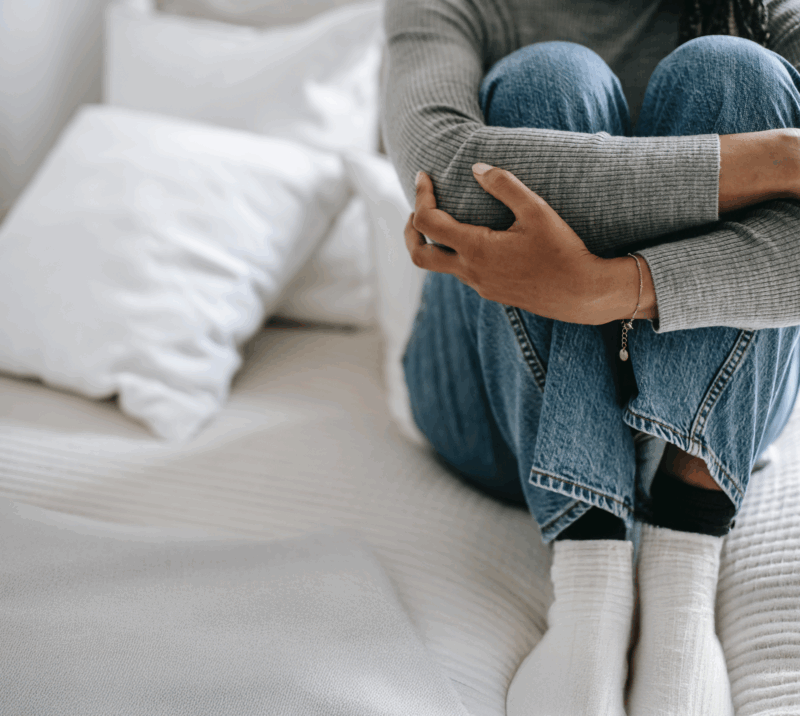 Young women sits curled up on a bed with white bedding. She had her arms folded over her legs, hugging them.