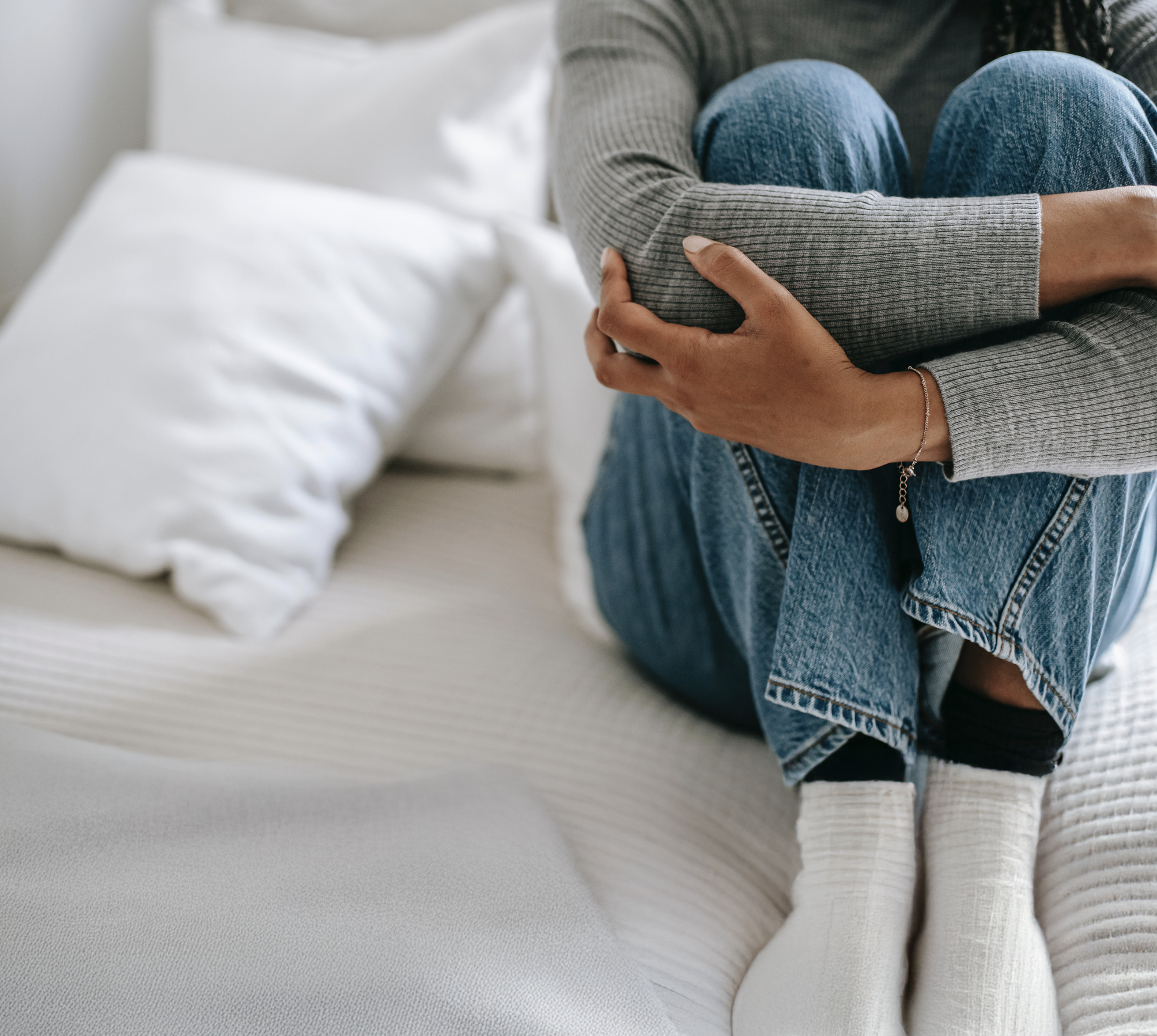 Young women sits curled up on a bed with white bedding. She had her arms folded over her legs, hugging them.