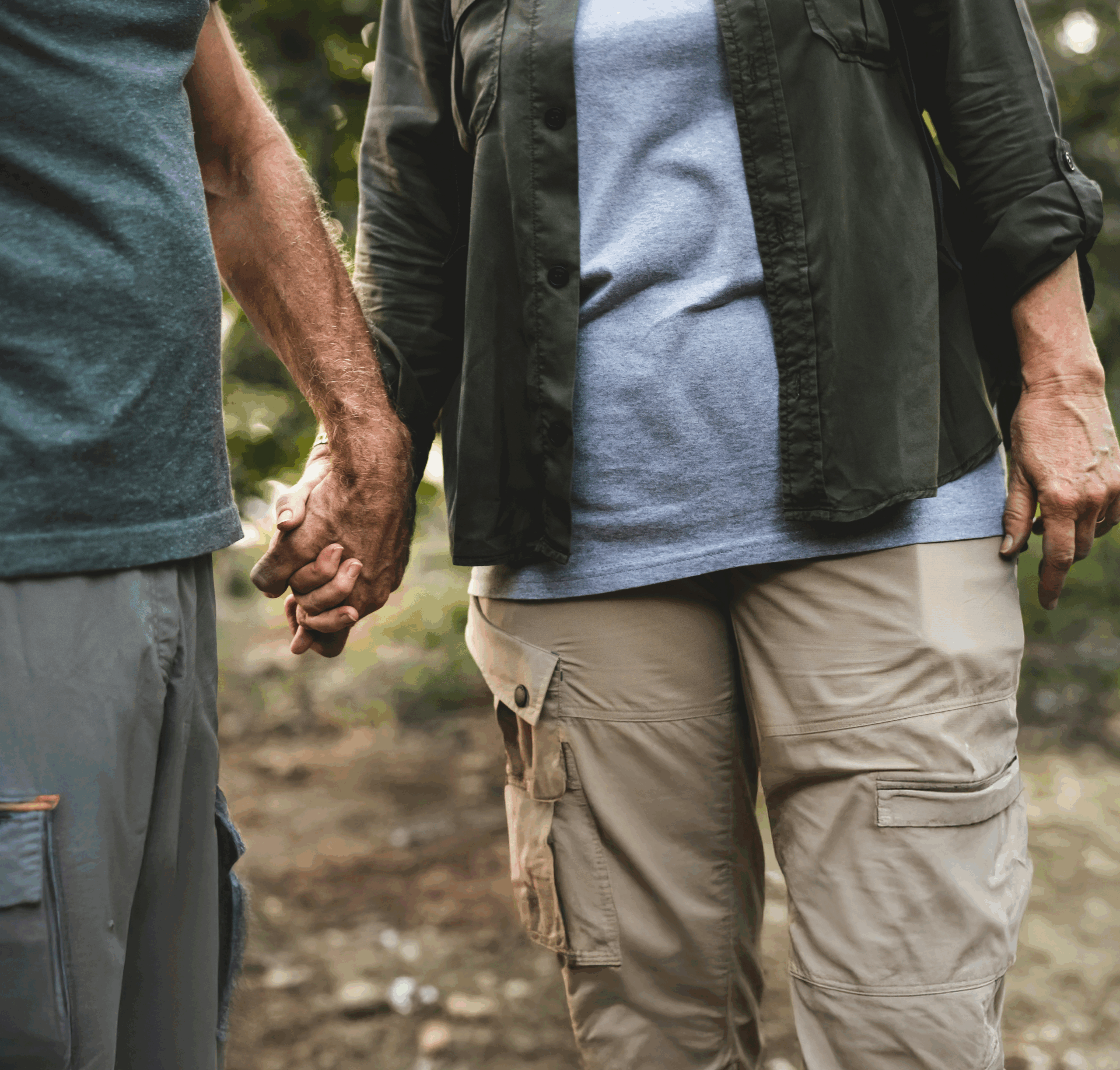 An older couple of unknown genders stand in front of a worn field track. The focus of the shot is their hand-holding.