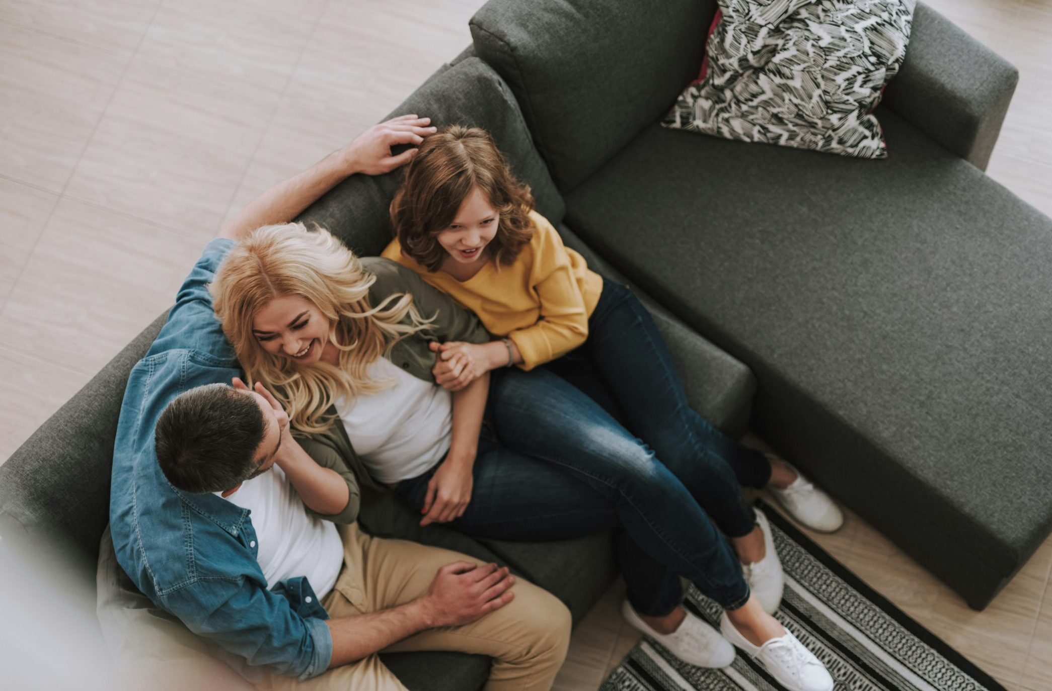 A family of three is sat on a dark grey corner sofa. The shot is from an arial view. They are laughing and smiling.