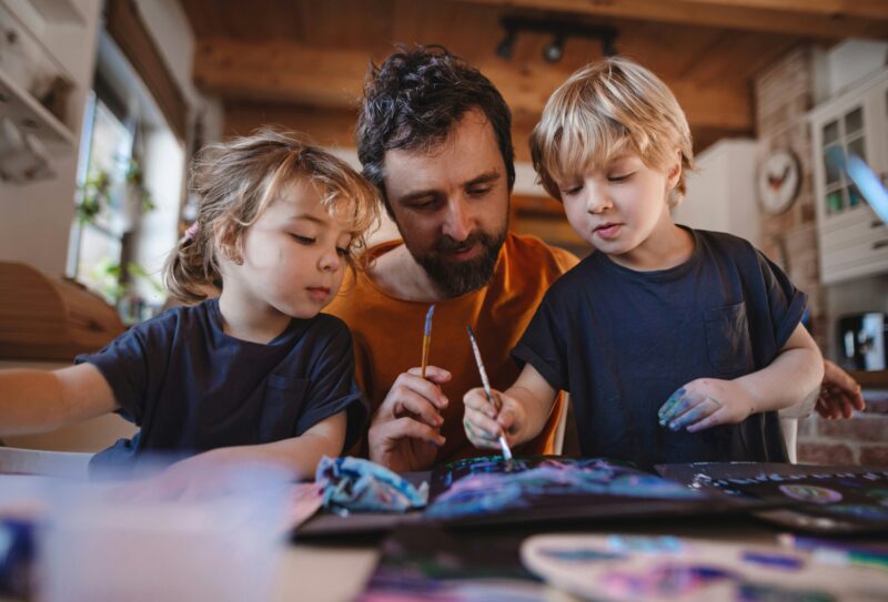 A father leans over a table where two blonde children are painting. They each have a brush in hand and paint marks on their face.