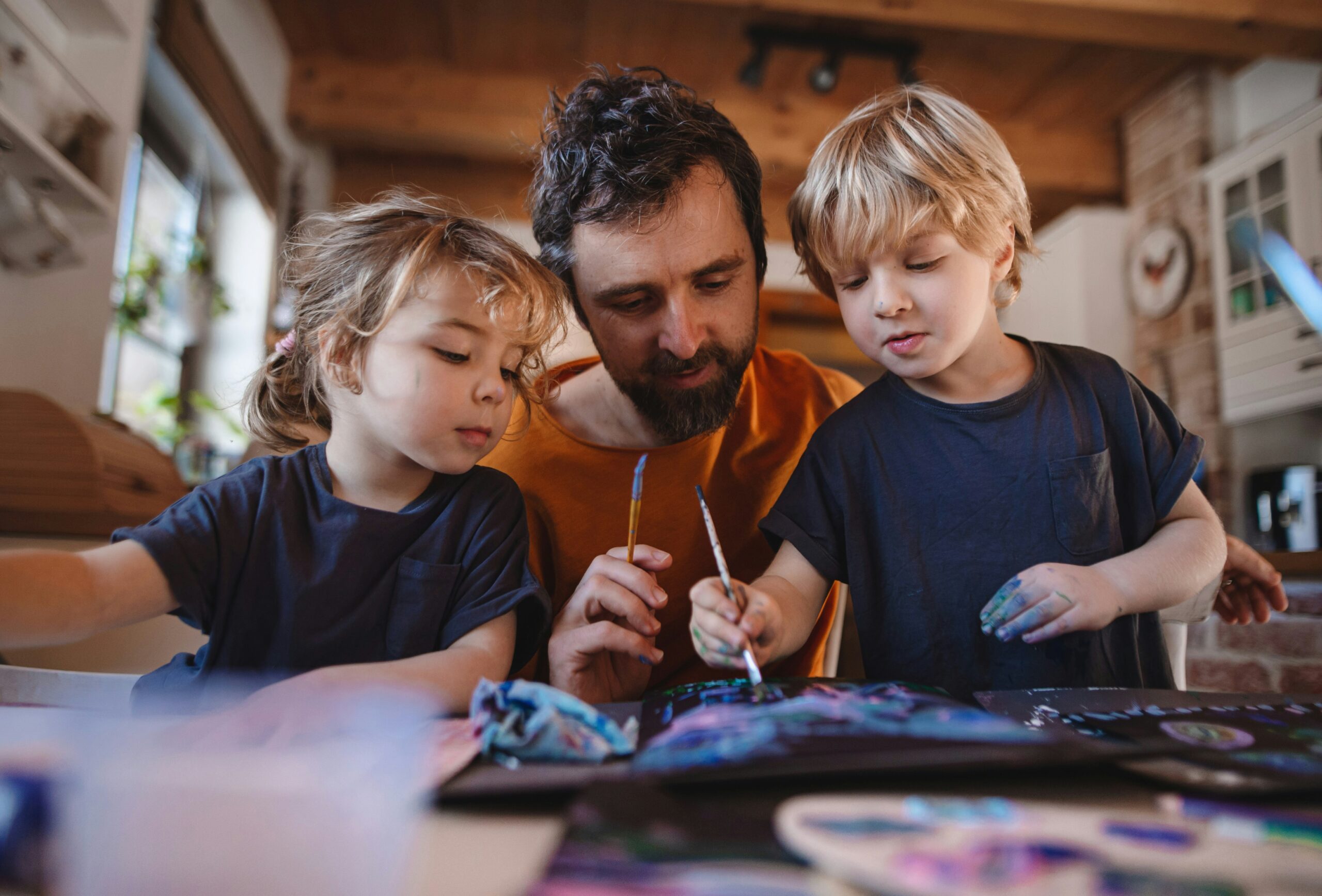 A father leans over a table where two blonde children are painting. They each have a brush in hand and paint marks on their face.