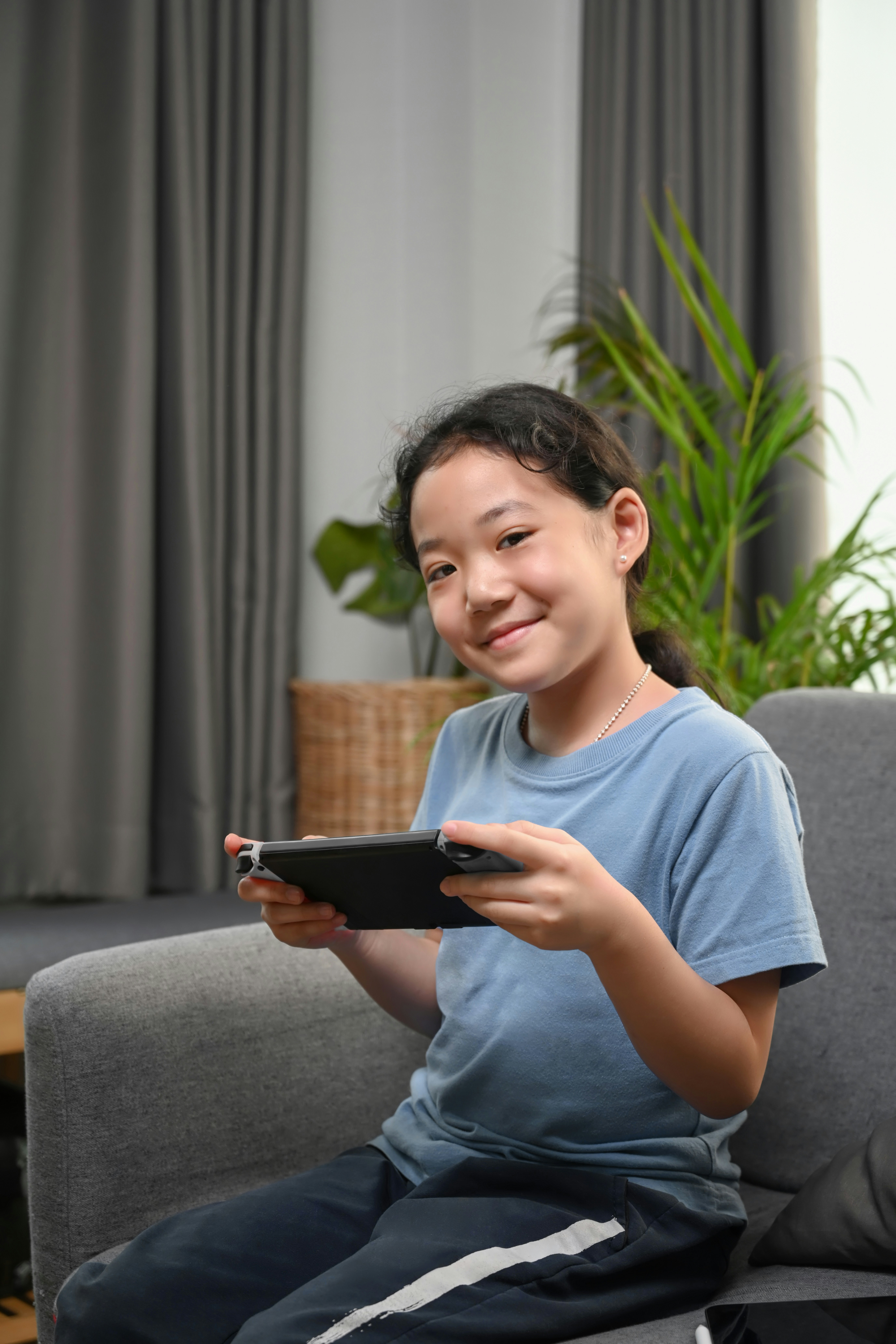 Young asian girl with dark hair and a light blue t-shirt sat on a couch smiling with a hand held gaming device