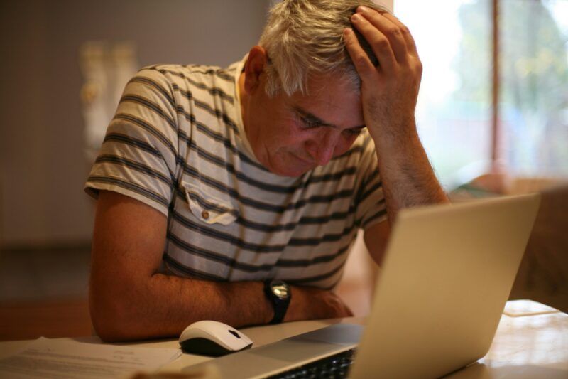 An older white man with white hair and a striped t-shirt looking down towards his laptop with a sad and drained looked.