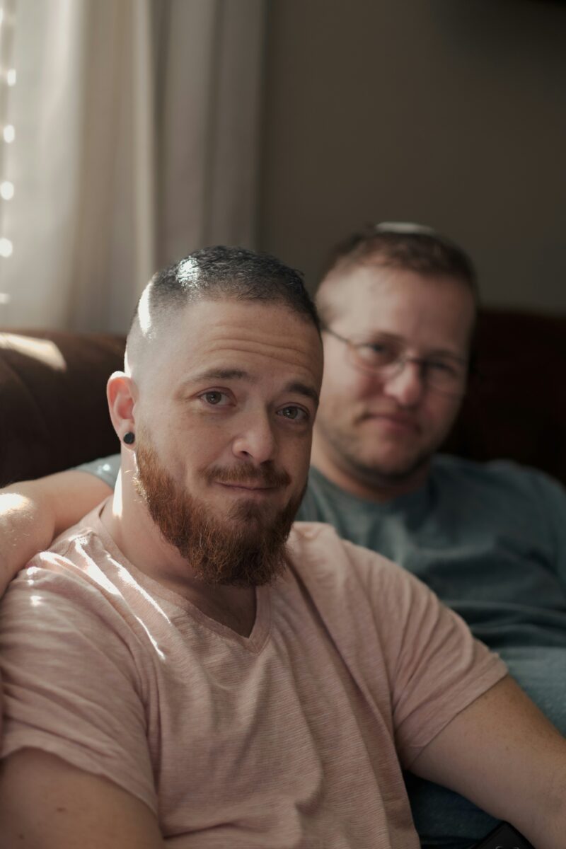 A male couple sat together one white man in the foreground with a pale beige t-shirt, short hair and a beard. In the background a white man with short blonde hair in the background and a blue t-shirt.