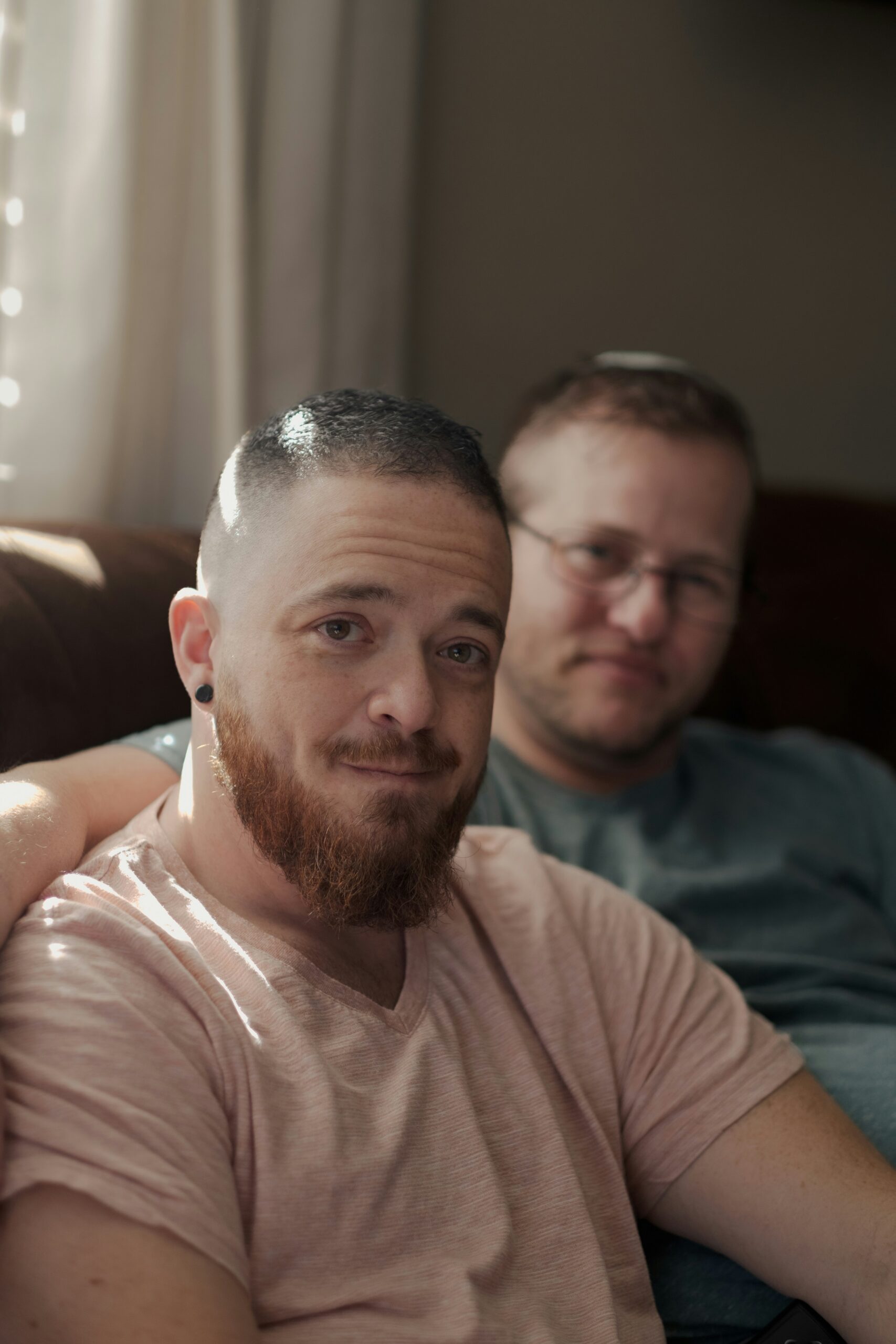 A male couple sat together one white man in the foreground with a pale beige t-shirt, short hair and a beard. In the background a white man with short blonde hair in the background and a blue t-shirt.