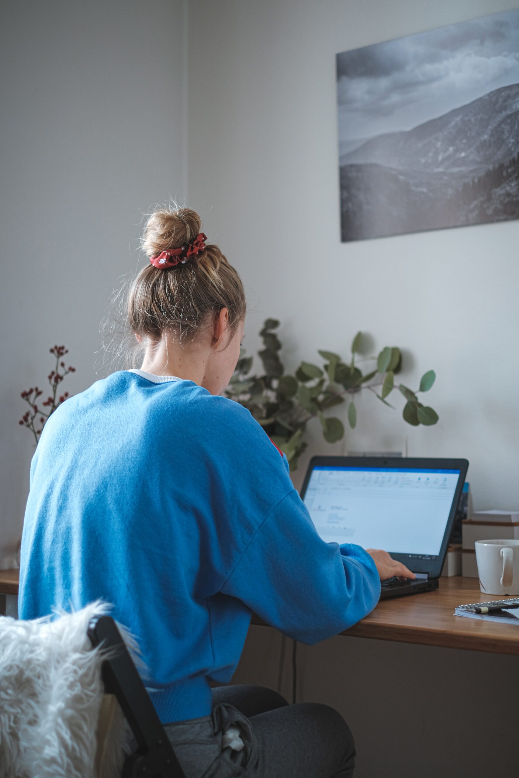 blonde woman sits at a desk and looks to be working from home in sweats and a scrunchie